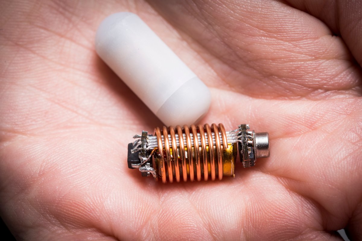 A swallowable sensor, silver wrapped in copper coil, held in a palm next to the white pill-like capsule used for ingestion.