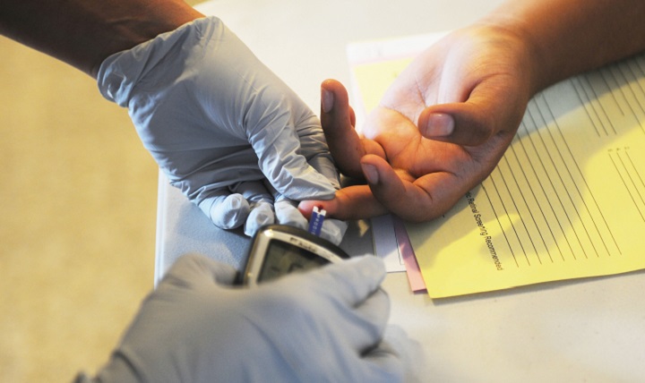 A pair of gloved hands testing blood sugar from a patient's hand using a finger-prick testing system. The discovery of viral insulin suggests viruses may have something to do with developing diabetes.