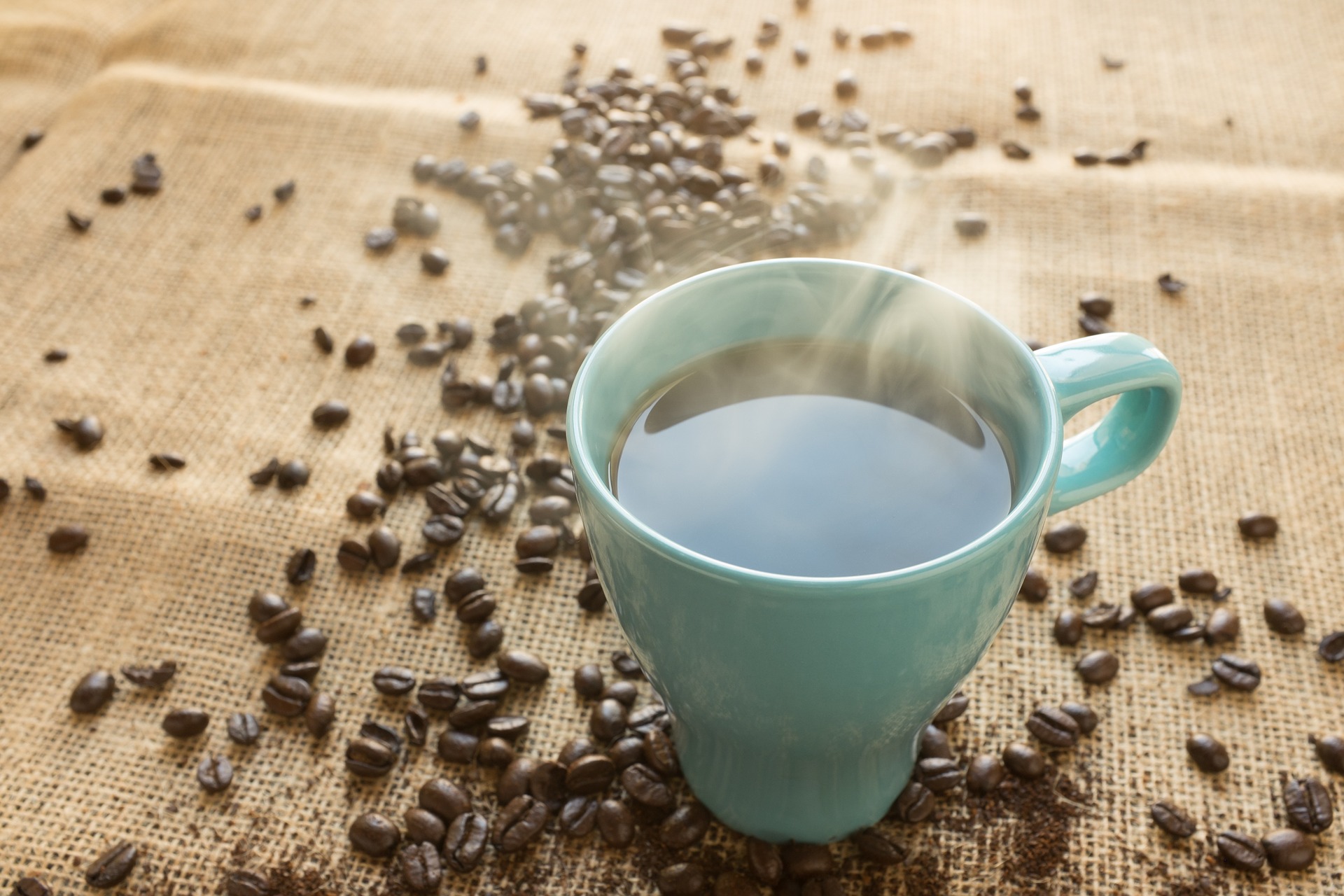A cup of coffee in a blue cup on a sheet of burlap scattered with coffee beans. The science over whether this cup or those beans increase your cancer risk is still shaky.