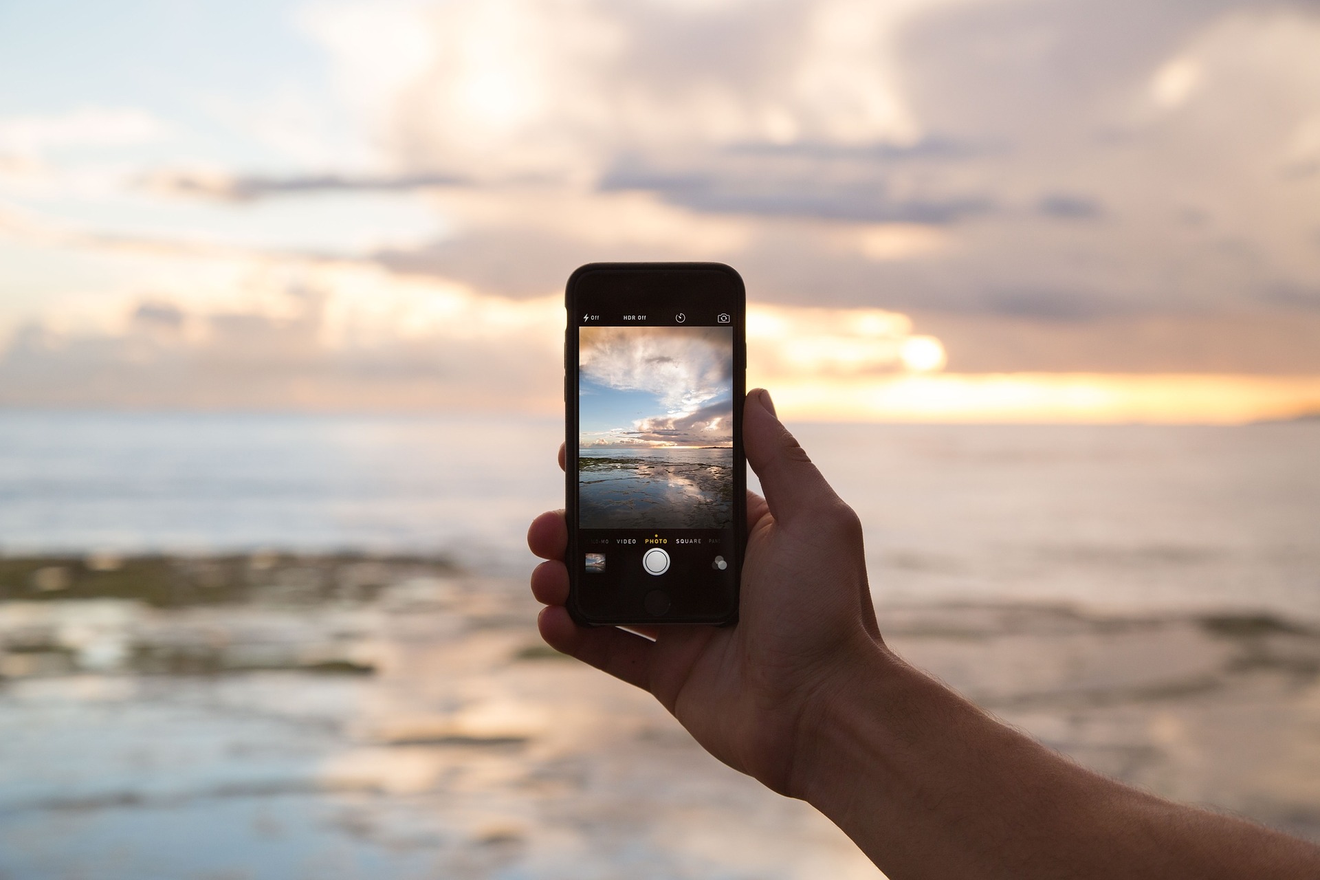 A hand holding a mobile phone taking a photo of a sunset over the ocean. Apps like Instagram encourage obsession with sharing content like this to the point that it may damage mental health.
