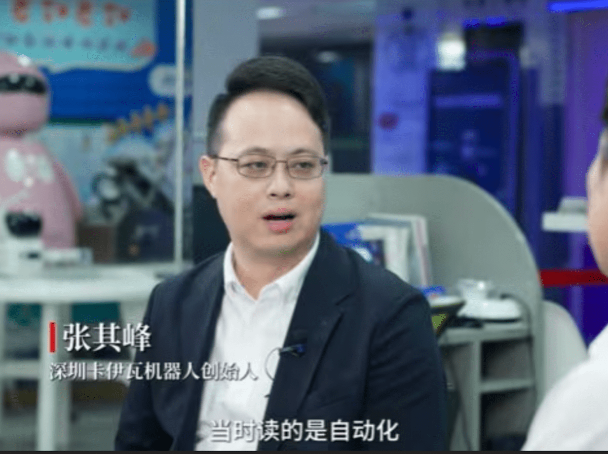An Asian man with round, wire-rimmed glasses looks at someone off camera in what appears to be a newsroom, which is bathed in blue and chrome. There is white text over the top of the image, written in Mandarin.