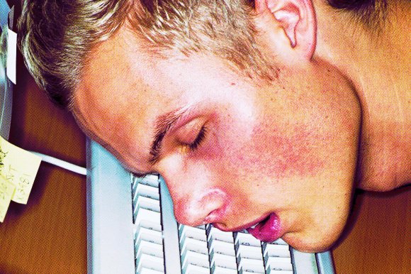 Close-up of a young person with light hair resting their face on a computer keyboard, eyes closed and mouth slightly open, suggesting exhaustion or sleep. The keyboard is white with visible keys, and there are sticky notes attached to the side of the desk. The skin has a slightly reddish tone.