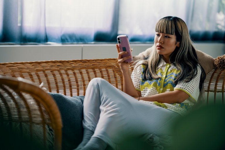 A woman with long, dark hair featuring blonde highlights and bangs is sitting on a wicker sofa. She is wearing a white crocheted top with yellow and blue patterns and light-colored pants. She is holding a pink smartphone and looking at it with a thoughtful expression. The background shows a window with sheer curtains.