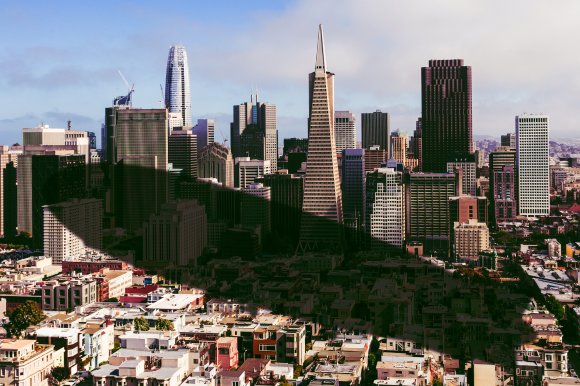 A city skyline featuring a mix of modern and older buildings under a partly cloudy sky. The shadow of an airship has been overlaid onto them.