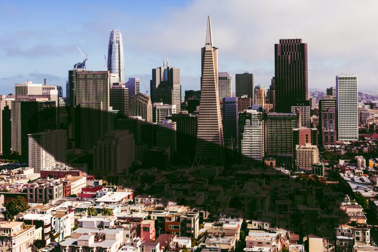 A city skyline featuring a mix of modern and older buildings under a partly cloudy sky. The shadow of an airship has been overlaid onto them.