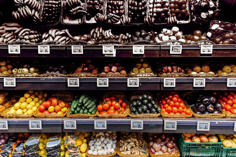 Shelves filled with a variety of fresh fruits and vegetables arranged in baskets, including bananas, tomatoes, cucumbers, lemons, eggplants, garlic, ginger, and onions, with price tags displayed below each section.