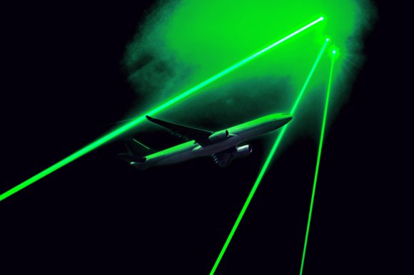 Illustration showing an airplane flying through a dark sky illuminated by multiple bright green laser beams. The lasers create a glowing effect around the aircraft, highlighting its wings and fuselage.