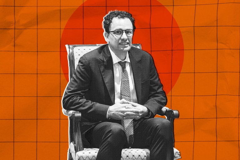 Dario Amodei is seated on an upholstered armchair. He is wearing a dark suit, a white shirt, and a patterned tie. His hands are clasped together in front of him. The background features an orange grid pattern with a large red circle behind him. The image has a stylized, graphic design effect.