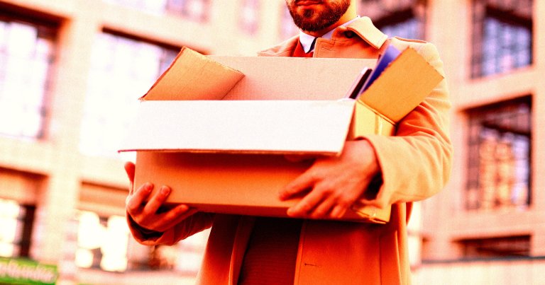 A person wearing a beige coat is holding an open cardboard box filled with various items, including a blue folder or notebook. The background shows a blurred urban setting with buildings and windows.