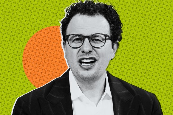 Anthropic CEO Dario Amodei is shown with curly dark hair and glasses is wearing a dark pinstripe suit jacket over a white shirt. He has a slightly open mouth expression. The background is a green grid pattern with an orange circle behind his head.