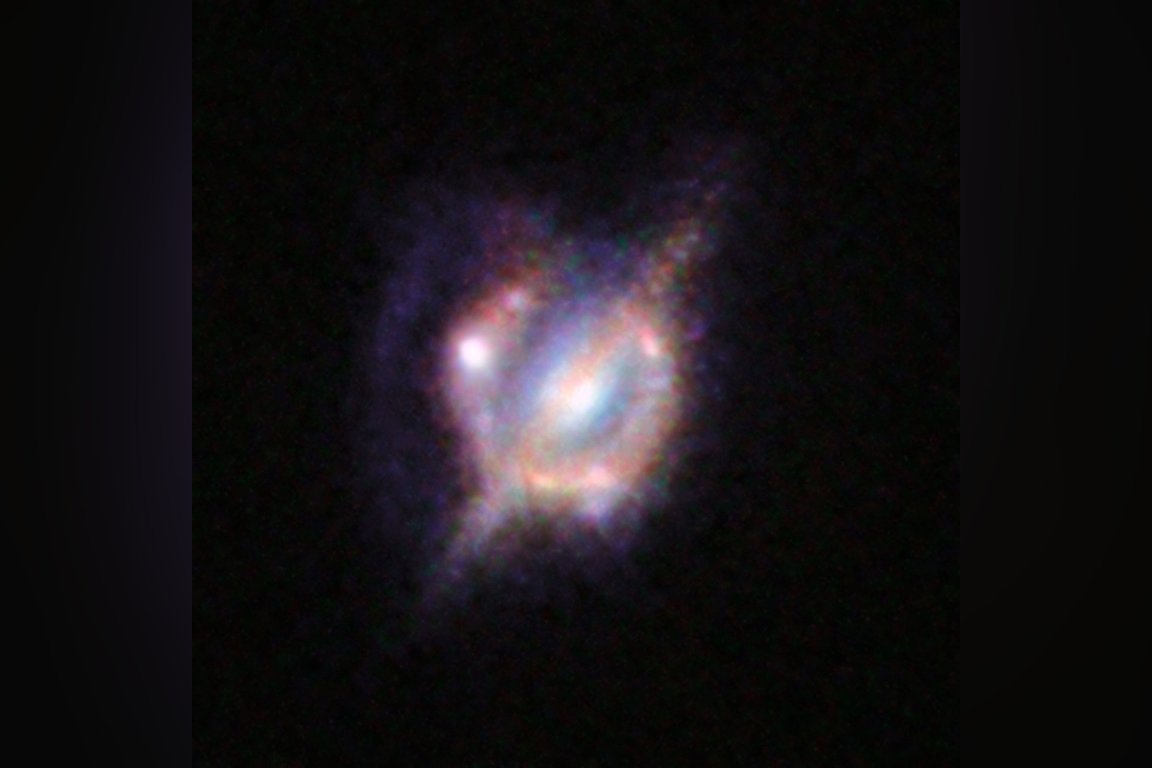 A colorful, irregularly shaped galaxy with a bright core and faint, diffuse outer regions against a black background. The galaxy displays a mix of blue, purple, and pink hues, with some areas appearing more concentrated and others more diffuse, suggesting active star formation and complex structure.