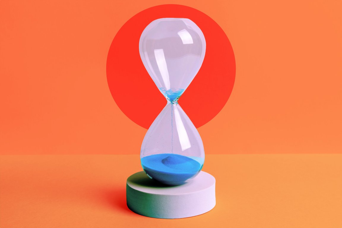 Transparent hourglass with blue sand flowing from the top to the bottom chamber, placed on a white circular pedestal against an orange background with a large red circle behind it.