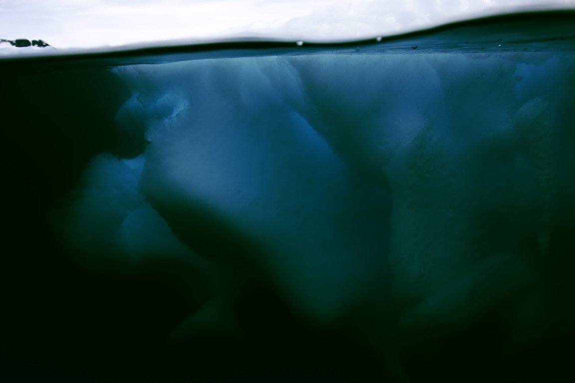 Underwater view of a large, dark blue iceberg with a smooth, rounded surface, partially submerged beneath a calm water surface with a cloudy sky above.