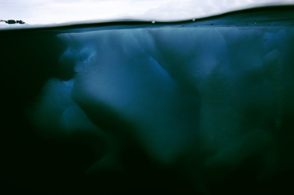Underwater view of a large, dark blue iceberg with a smooth, rounded surface, partially submerged beneath a calm water surface with a cloudy sky above.