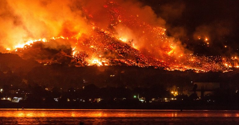 A large wildfire is burning intensely on a hillside at night, with bright orange flames and glowing embers spreading across the landscape. Thick smoke rises into the dark sky, and the fire's glow reflects on the water in the foreground. Dim lights from buildings or homes are visible at the base of the hill.