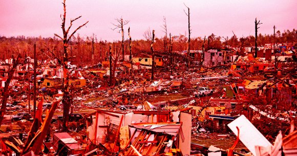 A devastated landscape showing extensive destruction of houses and trees, likely caused by a natural disaster such as a tornado or hurricane. The area is filled with debris, broken tree trunks, and remnants of buildings, with some vehicles partially buried under rubble. The sky is overcast, adding to the somber atmosphere.