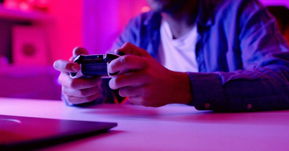 Hands holding a video game controller, with a person wearing a blue shirt and white t-shirt in the background, illuminated by purple and pink lighting. A laptop is partially visible on the table in front.