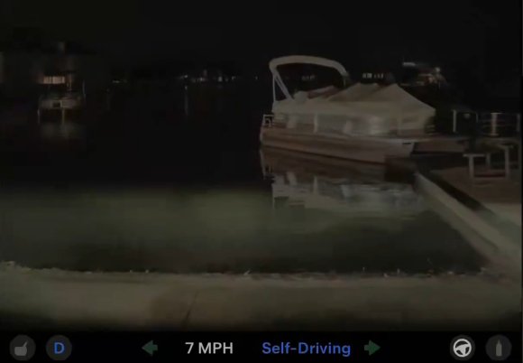 Still frame from video by Tesla driver Daniel Milligan. A boat docked at a pier at night, with its reflection visible in the calm water. The image is taken from a vehicle dashboard camera, showing a speed of 7 MPH and indicating that the vehicle is in self-driving mode.