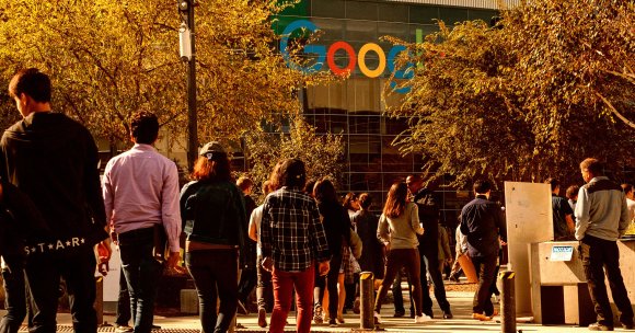 People walking toward a Google office building, surrounded by trees with autumn-colored leaves. The Google logo is prominently displayed on the glass façade of the building. The scene is lit by warm sunlight.