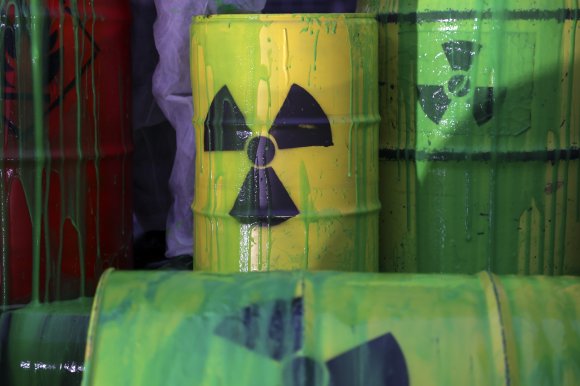 Yellow and green metal barrels with radioactive hazard symbols painted in black, covered in dripping green paint. A red barrel is partially visible on the left side, also with green paint dripping down. The barrels appear worn and stained.