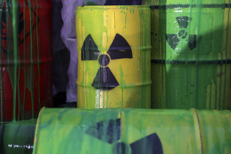 Yellow and green metal barrels with radioactive hazard symbols painted in black, covered in dripping green paint. A red barrel is partially visible on the left side, also with green paint dripping down. The barrels appear worn and stained.