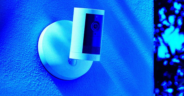 White Ring security camera mounted on a textured wall, illuminated with blue lighting. The camera has a cylindrical shape with a lens and sensor visible on the front.