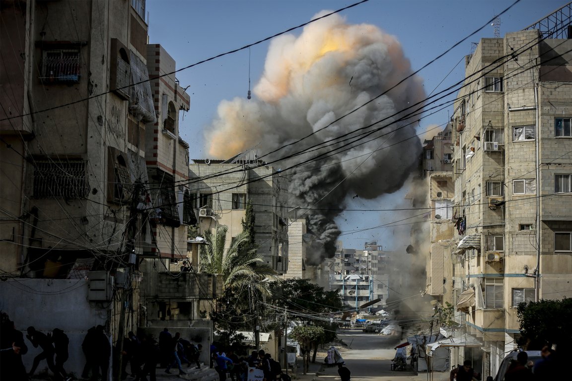 A large explosion with thick black and gray smoke billowing into the sky in an urban area. The explosion is centered between two multi-story buildings with numerous windows and balconies. Electrical wires crisscross the scene, and several people are visible on the street, some crouching or running away from the blast. The sky is clear and blue.