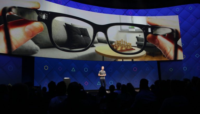Mark Zuckerberg stands on a stage in front of a large screen displaying a close-up image of black-framed glasses being held by two hands. Through the lenses, a clear view of a living room with a gray couch, black pillow, round wooden table, and a chessboard with pieces is visible, while the surrounding area outside the lenses appears blurred. The stage backdrop is blue with various geometric shapes in different colors.
