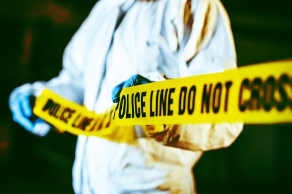 A person wearing a white protective suit and blue gloves is holding yellow police tape that reads "POLICE LINE DO NOT CROSS." The background is dark and out of focus.