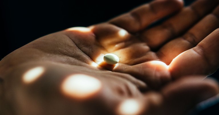 A close-up of an open hand with a single white pill resting in the center of the palm, illuminated by soft, scattered light spots against a dark background.