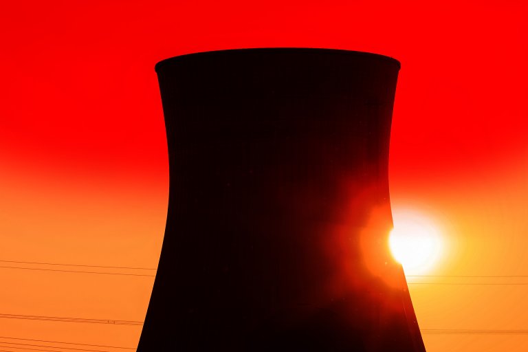 A large cooling tower silhouetted against a vibrant red and orange sunset sky, with the sun partially visible near the base of the tower. Thin power lines run horizontally across the lower part of the image.