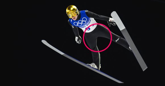Ski jumper in mid-air wearing a gold helmet, reflective goggles, black suit, and a blue and white bib with Olympic rings, against a black background. The skis are wide and angled, with one ski slightly raised.