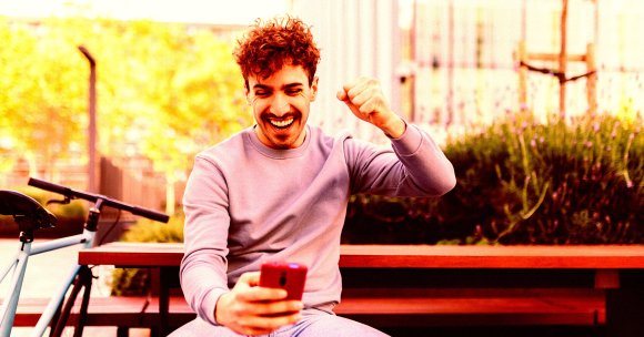 A young man with curly hair and a mustache is sitting on a wooden bench outdoors, smiling broadly and raising his fist in celebration while looking at a red smartphone in his hand. A light blue bicycle is parked next to the bench, and there are green bushes and a fence in the background.