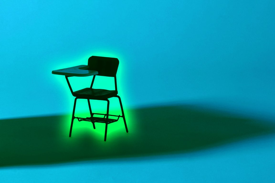 A black school desk chair with an attached writing surface is illuminated from behind by a green light, casting a dark shadow on a blue background.