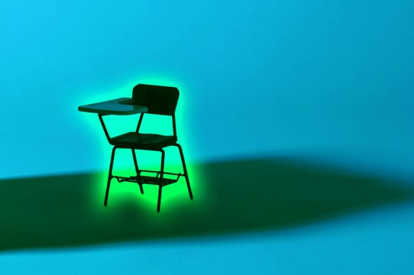 A black school desk chair with an attached writing surface is illuminated from behind by a green light, casting a dark shadow on a blue background.
