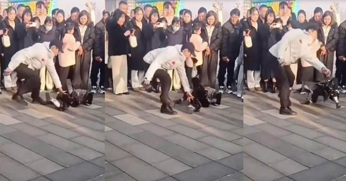 A man in a white jacket and black pants is interacting with a robot on a tiled outdoor surface. The robot has kicked the man in the nose while a crowd of people stands in the background watching and taking photos or videos.