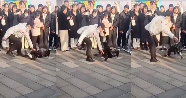 A man in a white jacket and black pants is interacting with a robot on a tiled outdoor surface. The robot has kicked the man in the nose while a crowd of people stands in the background watching and taking photos or videos.