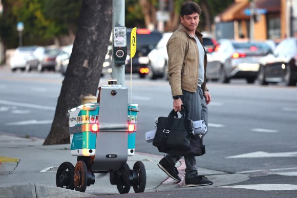 A four-wheeled delivery robot with a teal and gray body, equipped with lights and a yellow flag, is on a sidewalk near a pedestrian crossing button. A man wearing a brown jacket, gray shirt, and striped pants is walking nearby, carrying a black bag and a rolled-up mat. Cars are visible in the background on the street.