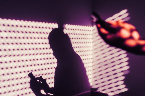 Shadow of a person holding and using a smartphone projected on a purple wall with a pattern of light dots. The shadow shows the person's head and hand clearly, with a blurred hand holding the phone in the foreground.
