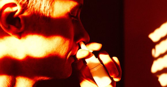A close-up of a person using a nasal spray, with warm, orange-red lighting casting shadows across their face and hand. The person is holding the white nasal spray bottle up to one nostril.