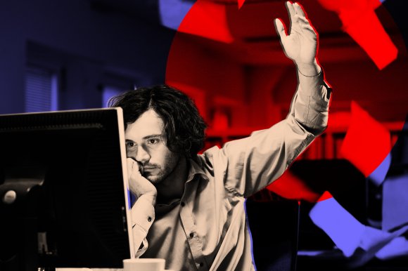 A man with curly hair and a beard sits at a desk, resting his chin on his hand while looking at a computer monitor. He raises his other hand, appearing frustrated or overwhelmed. Red and blue lighting effects highlight the background and scattered papers around him.