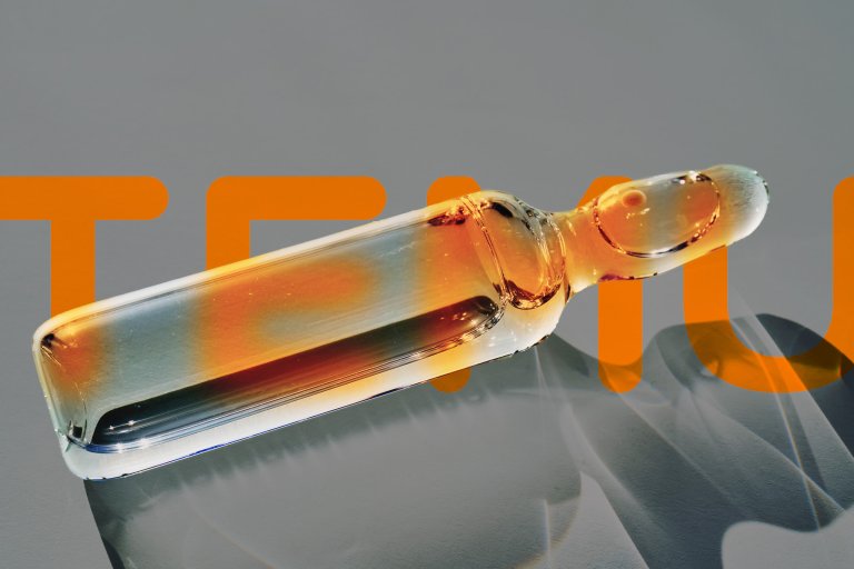 A close-up of a transparent glass ampoule filled with an amber-colored liquid, lying on a gray surface with an orange text partially visible in the background. The ampoule casts a shadow on the surface.