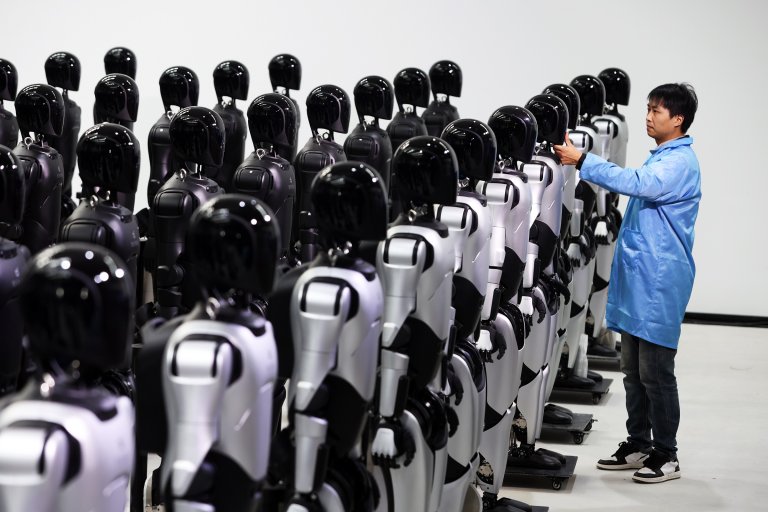 A man in a blue lab coat is inspecting or adjusting one of many humanoid AgiBot robots arranged in rows. The robots have black, glossy heads and silver and black bodies, standing on platforms with wheels. The background is plain white, emphasizing the robots and the man.