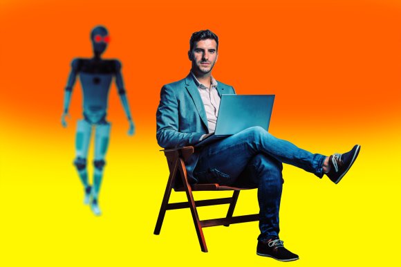 Man in a gray blazer and jeans sitting on a wooden chair with a laptop on his lap, against a gradient orange to yellow background. A humanoid robot with glowing red eyes is blurred in the background.