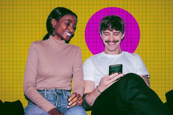 A smiling woman wearing a beige turtleneck and blue jeans sits next to a smiling man with a mustache, white t-shirt, and black pants who is looking at a smartphone. The background features a yellow grid pattern with a large purple circle behind the man.