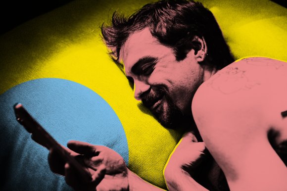 A man with a beard and mustache is lying on a yellow and blue textured surface, looking at a smartphone in his hand. He has short, slightly messy hair and multiple ear piercings. His skin and hair are digitally altered with a pink hue, and he has a tattoo on his upper arm. The image has a stylized, high-contrast color effect.