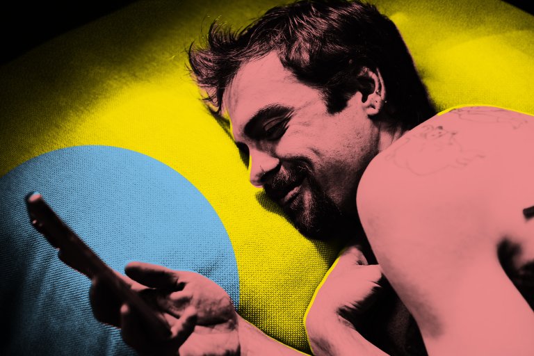 A man with a beard and mustache is lying on a yellow and blue textured surface, looking at a smartphone in his hand. He has short, slightly messy hair and multiple ear piercings. His skin and hair are digitally altered with a pink hue, and he has a tattoo on his upper arm. The image has a stylized, high-contrast color effect.