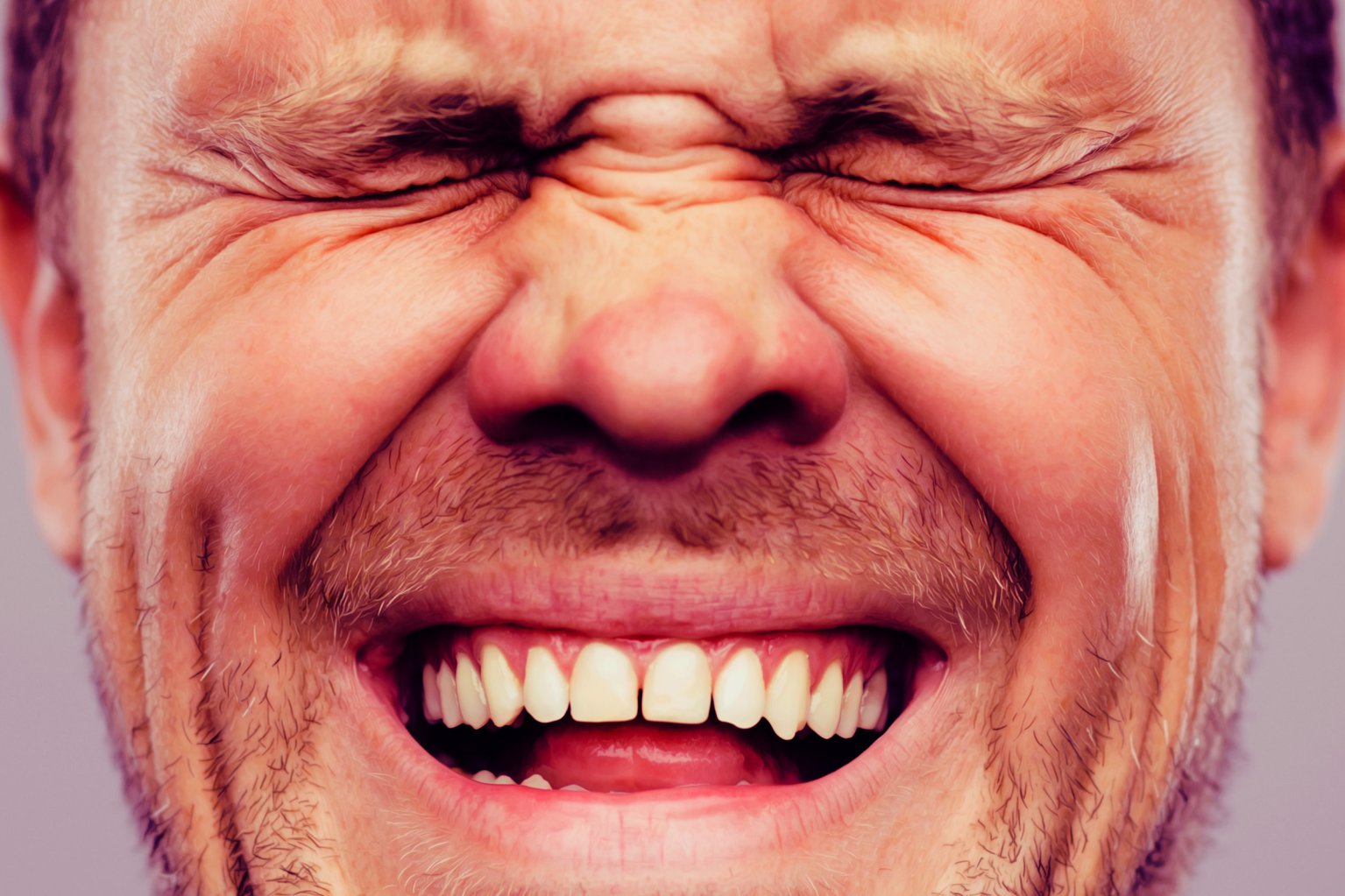 Close-up of a man's face with eyes tightly closed and a wide smile, showing his teeth and some facial stubble.