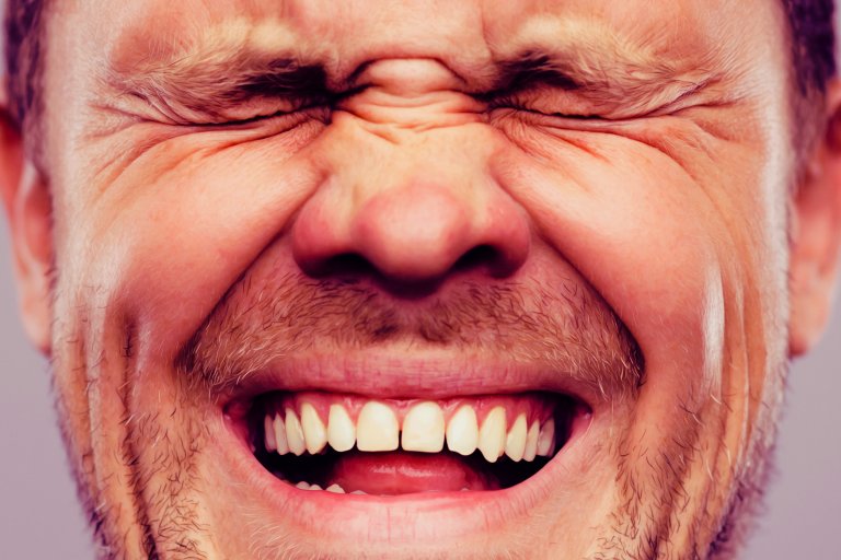 Close-up of a man's face with eyes tightly closed and a wide smile, showing his teeth and some facial stubble.