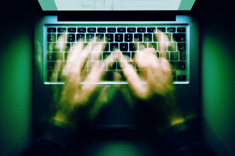 Hands typing rapidly on a laptop keyboard, captured with motion blur to emphasize speed. The laptop is viewed from above, with a faintly visible screen at the top. The overall lighting has a greenish tint.